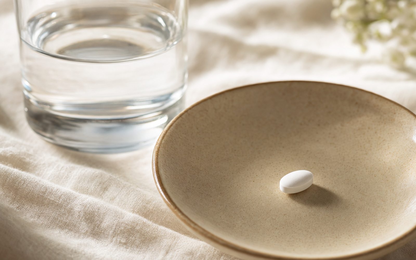 Editorial close-up of a single oval pill in a ceramic dish next to a glass of water — Foundayo oral GLP-1